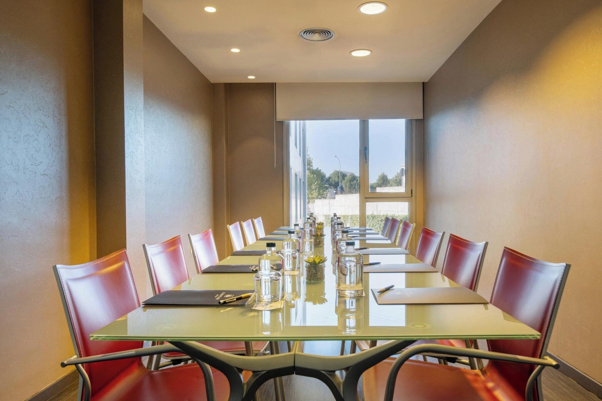 A conference room with a long table, red chairs, and a view of the outdoors through large windows.