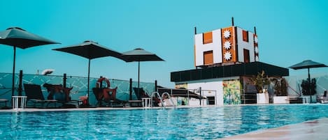 A swimming pool area with lounge chairs and umbrellas, a modern building with a distinctive facade, and a clear blue sky.