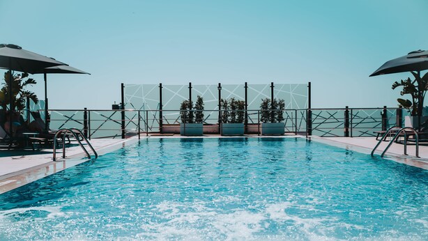 A swimming pool with clear blue water, surrounded by a metal fence and shaded by umbrellas.