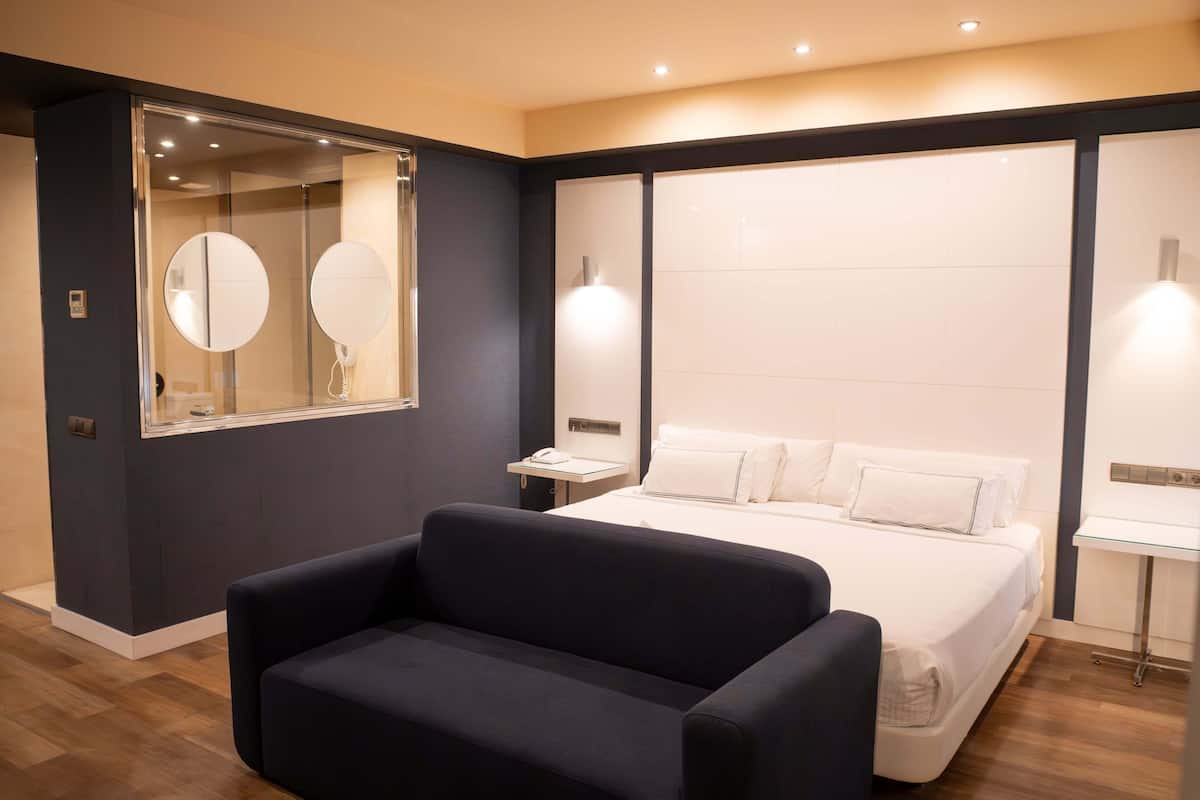 A modern hotel room with a large bed, a dark-colored sofa, a large mirror, and a bedside table with a lamp.