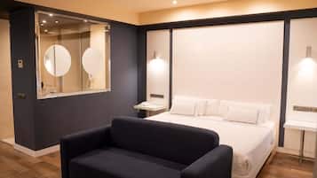 A modern hotel room with a large bed, a dark-colored sofa, a large mirror, and a bedside table with a lamp.