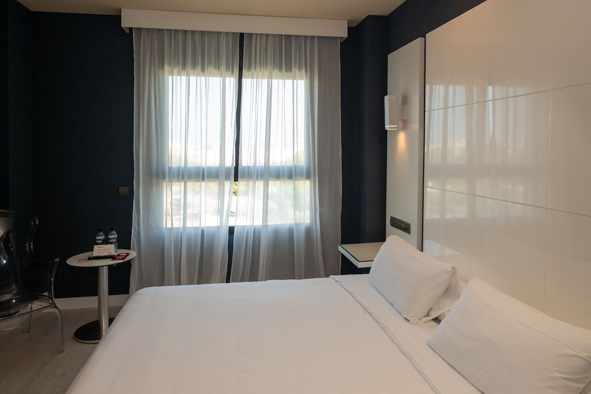 A hotel room with a large bed, a small table with a chair, a window with curtains, and a mirror on the wall.