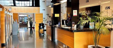 A modern hotel lobby with a reception desk, a large 'MN' sign, and a potted plant.