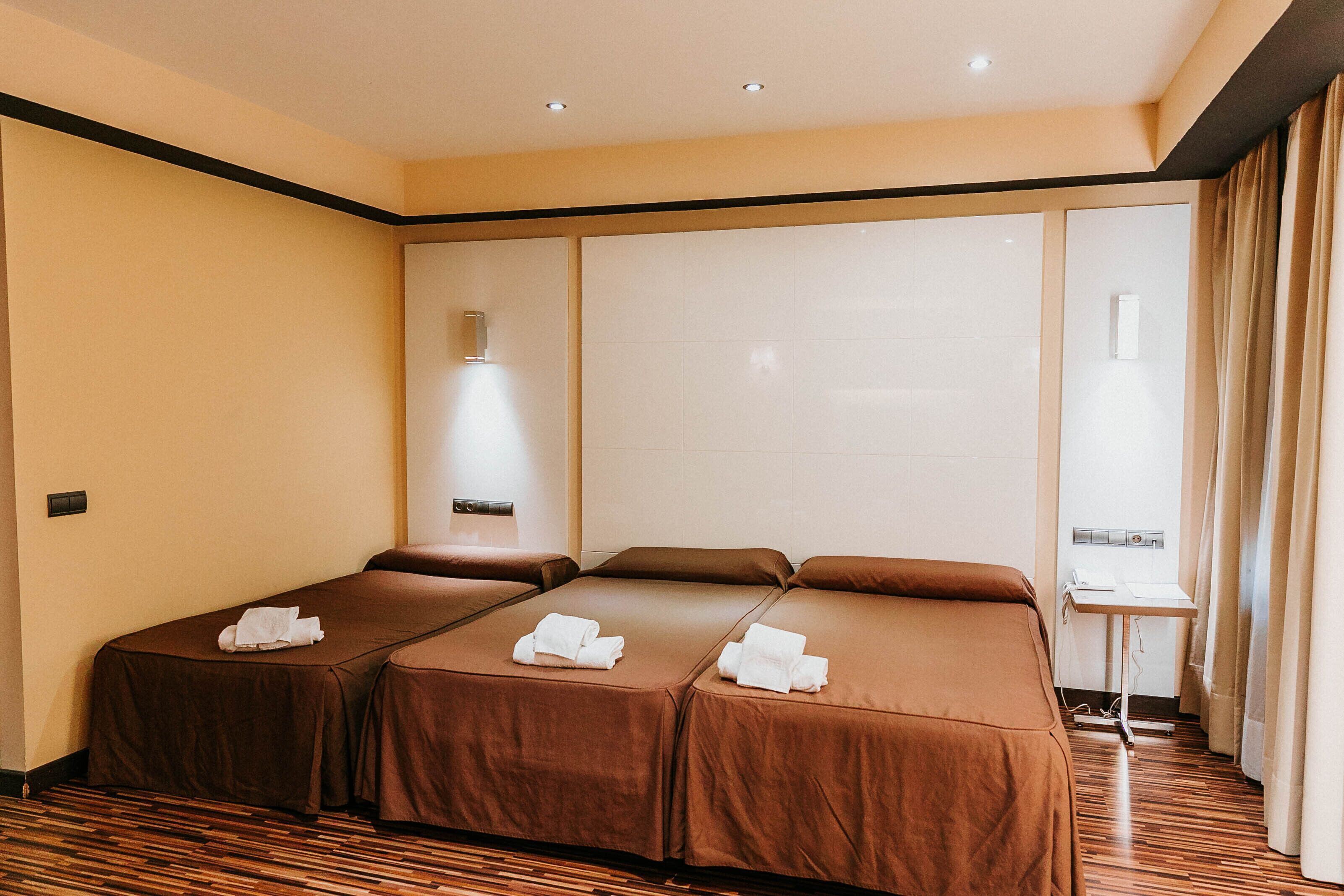 Quarto de hotel com duas camas, uma mesinha e uma janela com cortinas.