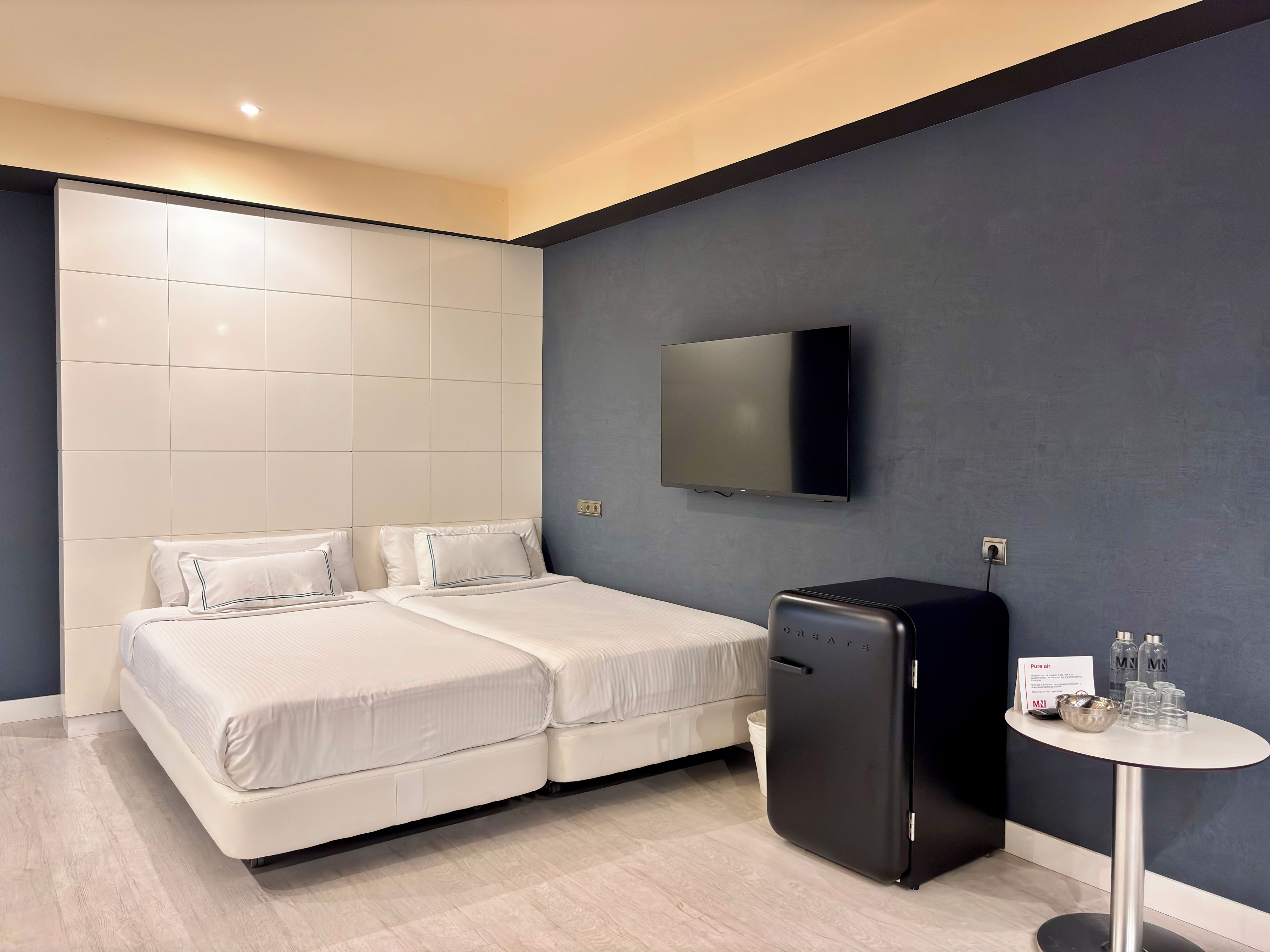 A modern hotel room with a bed, a flat-screen TV, a minibar, and a small table with glasses and bottles.