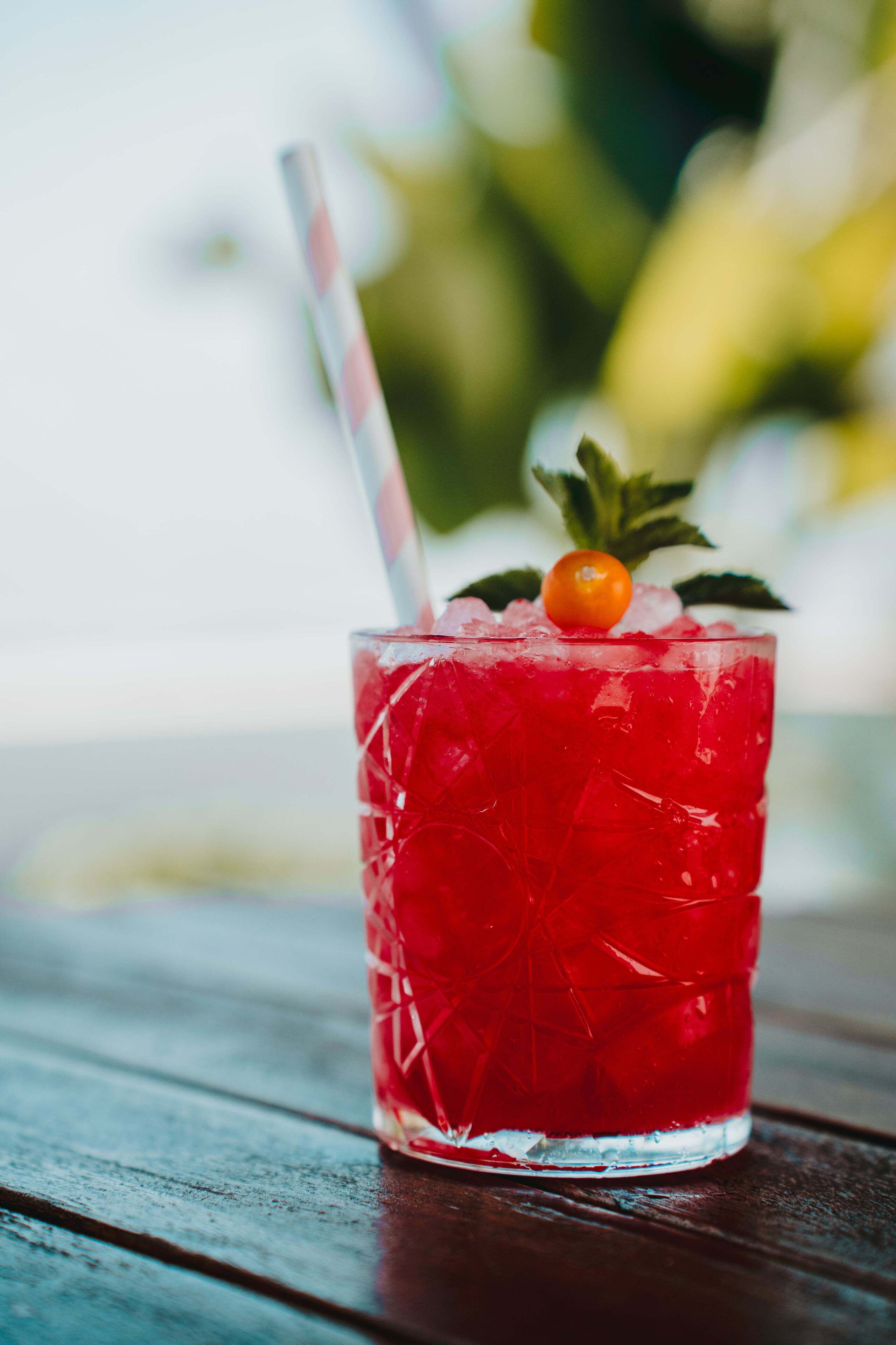 A glass with a red beverage, ice, a straw, and a cherry on top, placed on a wooden surface.
