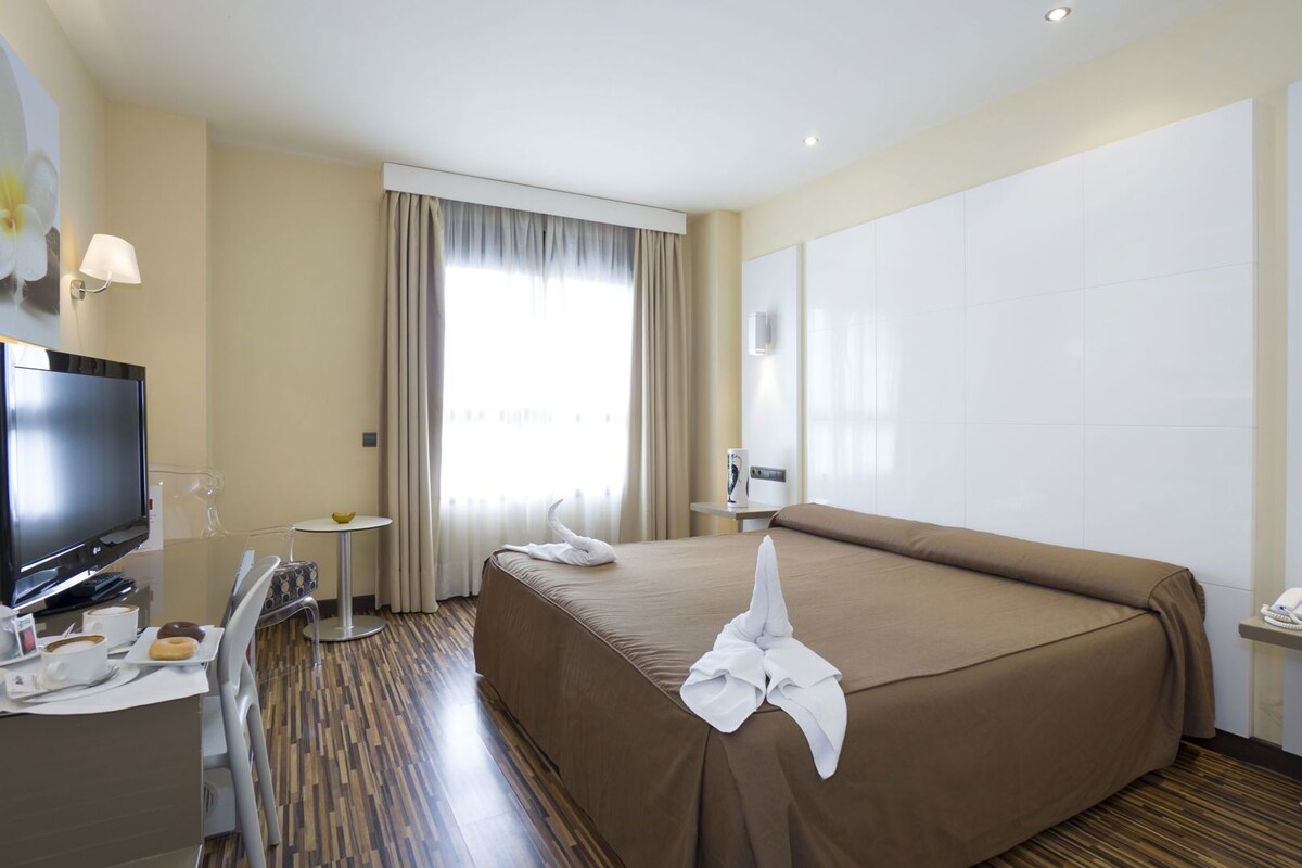 A hotel room with a bed, a TV, a desk with a phone, a coffee maker, and a window with curtains.