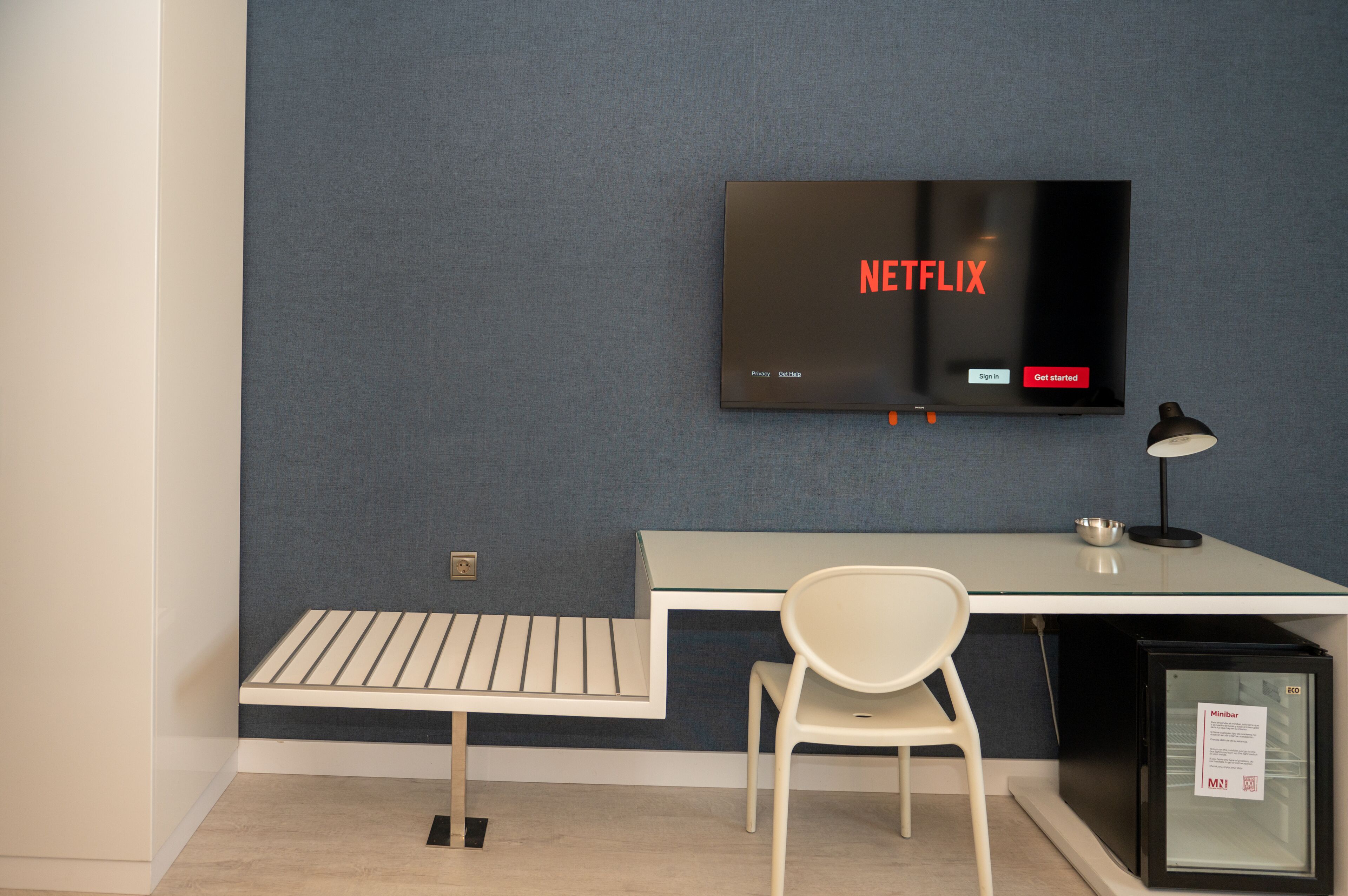 Een moderne werkruimte met een flatscreen waarop het Netflix logo staat, een wit bureau, een witte stoel en een kleine zwarte koelkast.