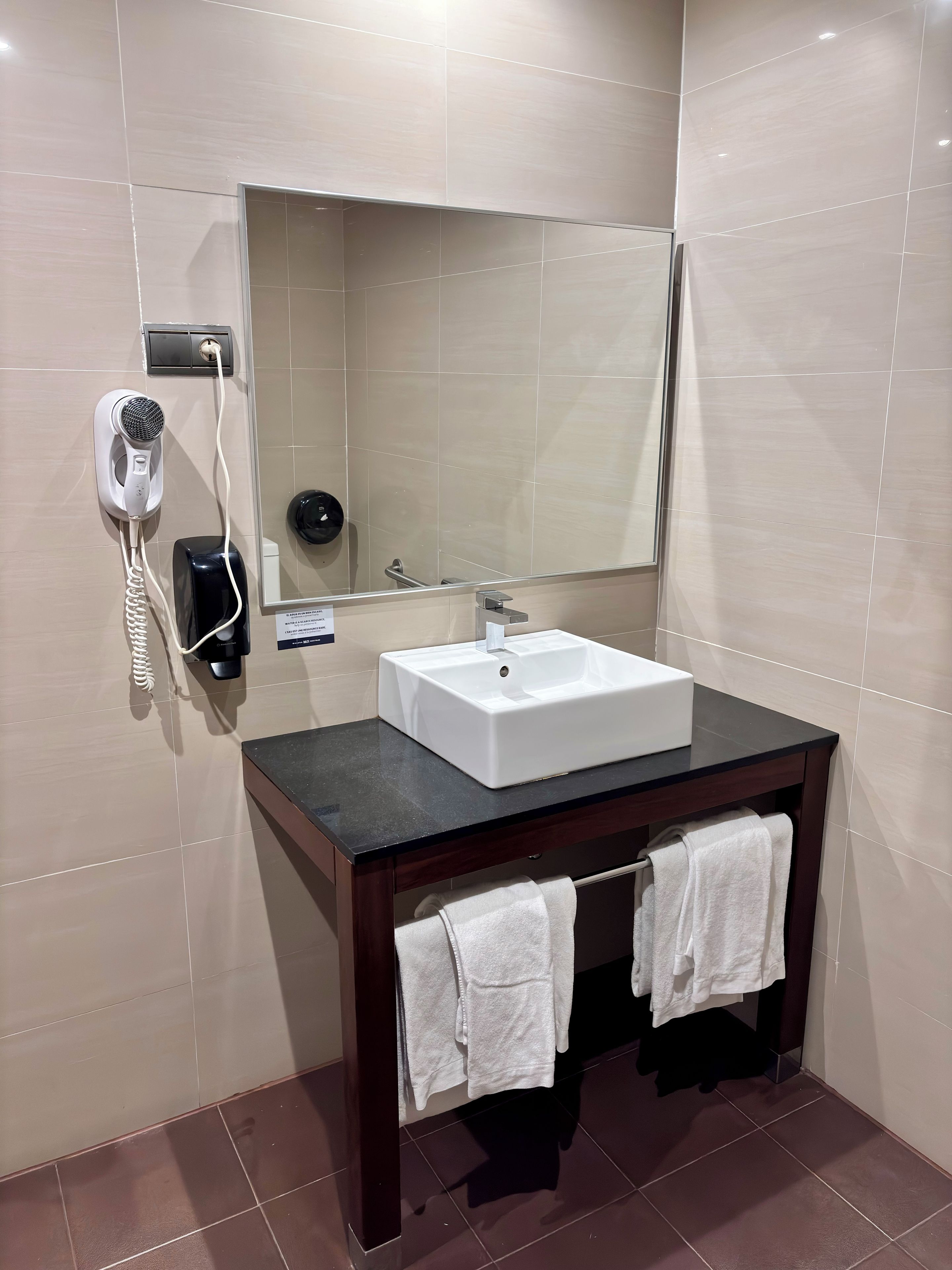 Un baño moderno con un lavabo blanco, encimera oscura y dos toallas colgadas en un perchero.