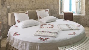 A bedroom with a stone wall, a bed with floral bedding, a bench, and a window with curtains. - Alacati Degirmen Otel (Cesme)