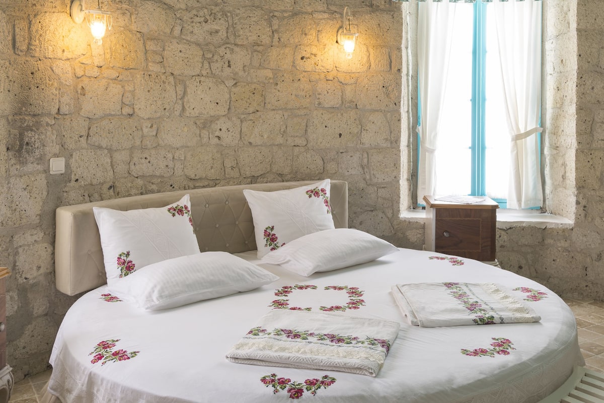A bedroom with a stone wall, a bed with floral bedding, a bench, and a window with curtains.
