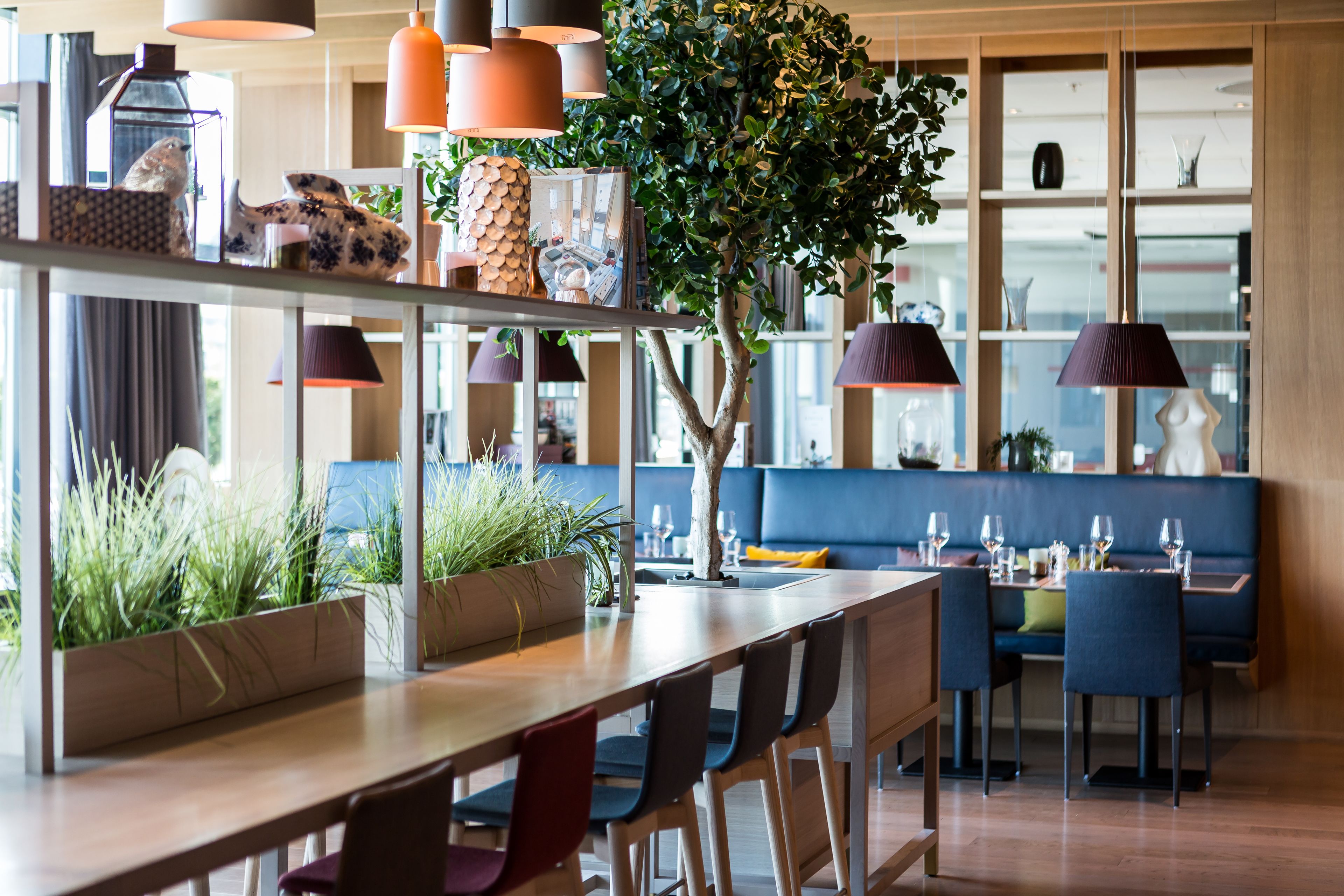 A modern dining area with a long wooden table, multiple chairs, and a blue booth. There are hanging pendant lights, a potted plant, and a bookshelf with various items.