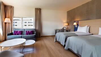 A hotel room with a sofa, two beds, a desk, and a view of a building.