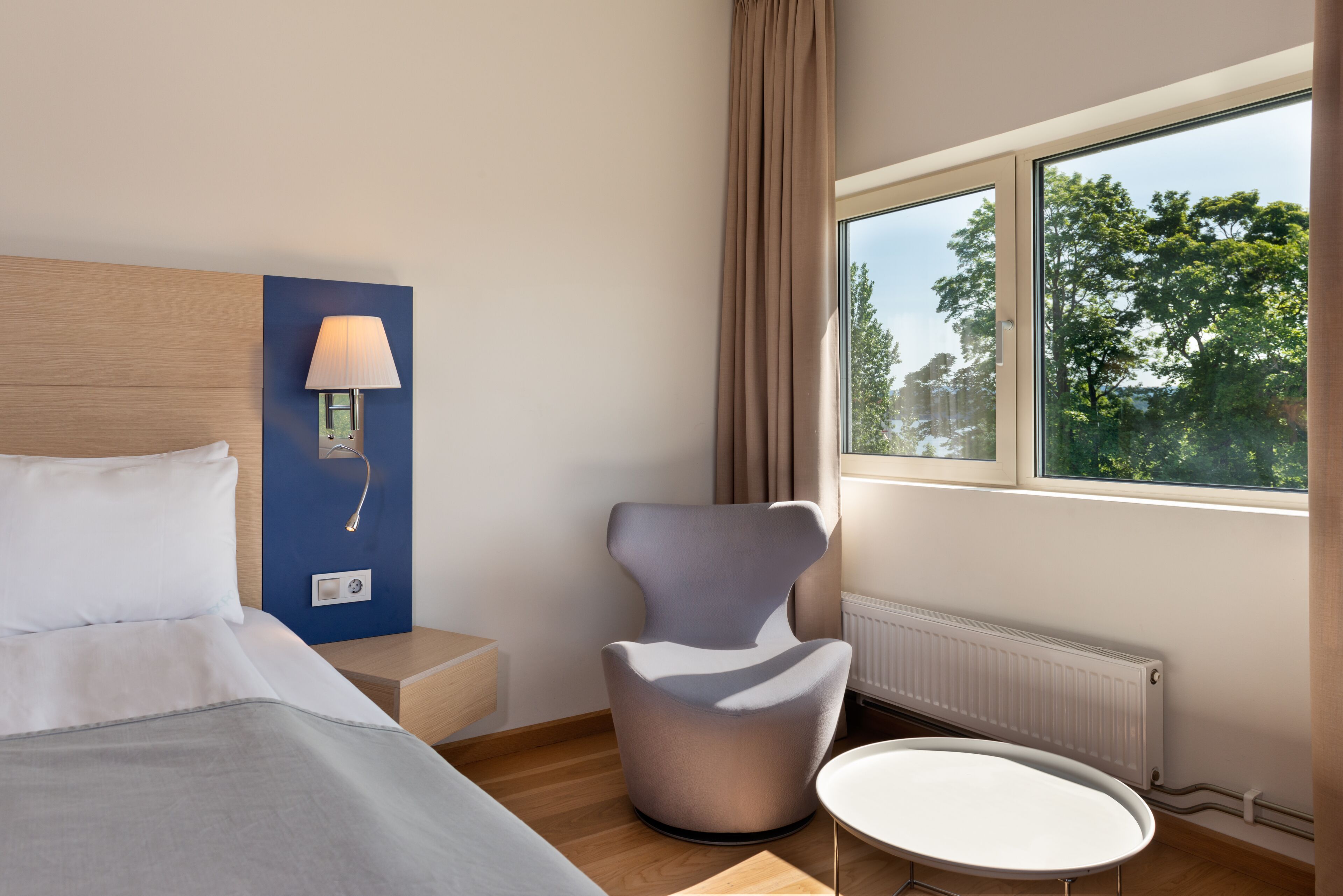 A modern hotel room with a bed, a chair, a small table, a lamp, and a window with a view of trees.