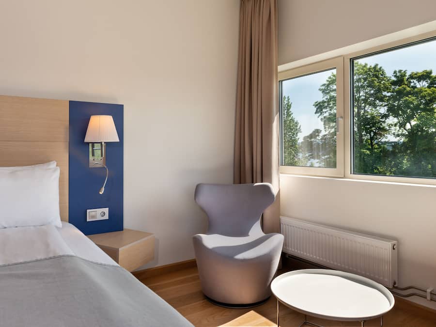 A modern hotel room with a bed, a chair, a small table, a lamp, and a window with a view of trees.