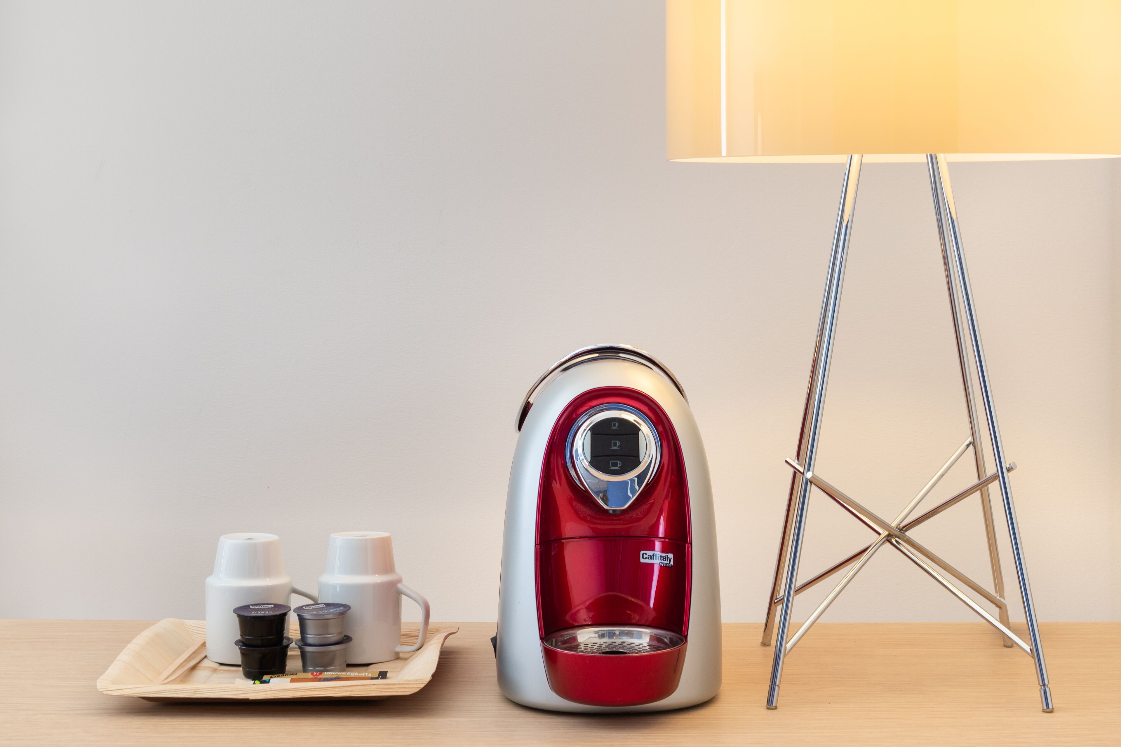 A red and silver electric kettle on a tray with two white cups and a small container, next to a lamp and a metal stand.