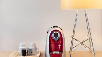 Una tetera eléctrica roja y plateada sobre una bandeja con dos tazas blancas y un pequeño recipiente, junto a una lámpara y un soporte metálico.