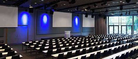 Un salón de conferencias con filas de mesas y sillas, un atril y pantallas grandes.