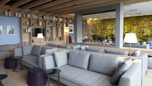 A modern living area with a grey sofa, purple ottomans, a flat-screen TV, bookshelves, and a green wall with plants.