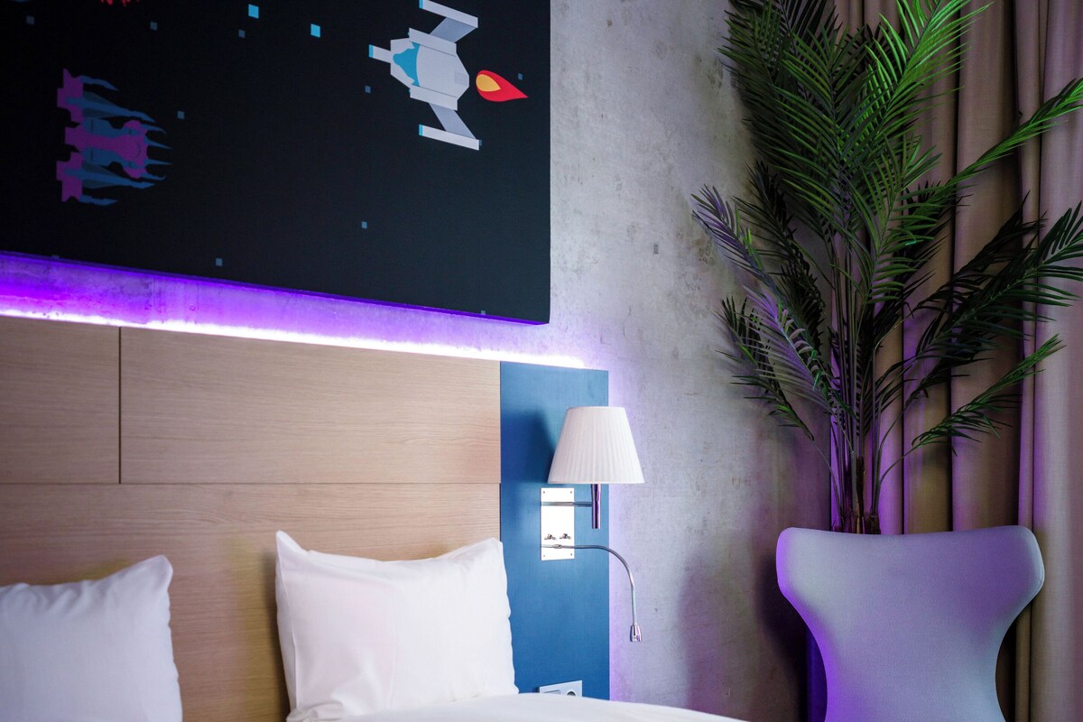 A hotel room with a bed, a nightstand, a wall-mounted lamp, a large potted plant, and a space-themed artwork.