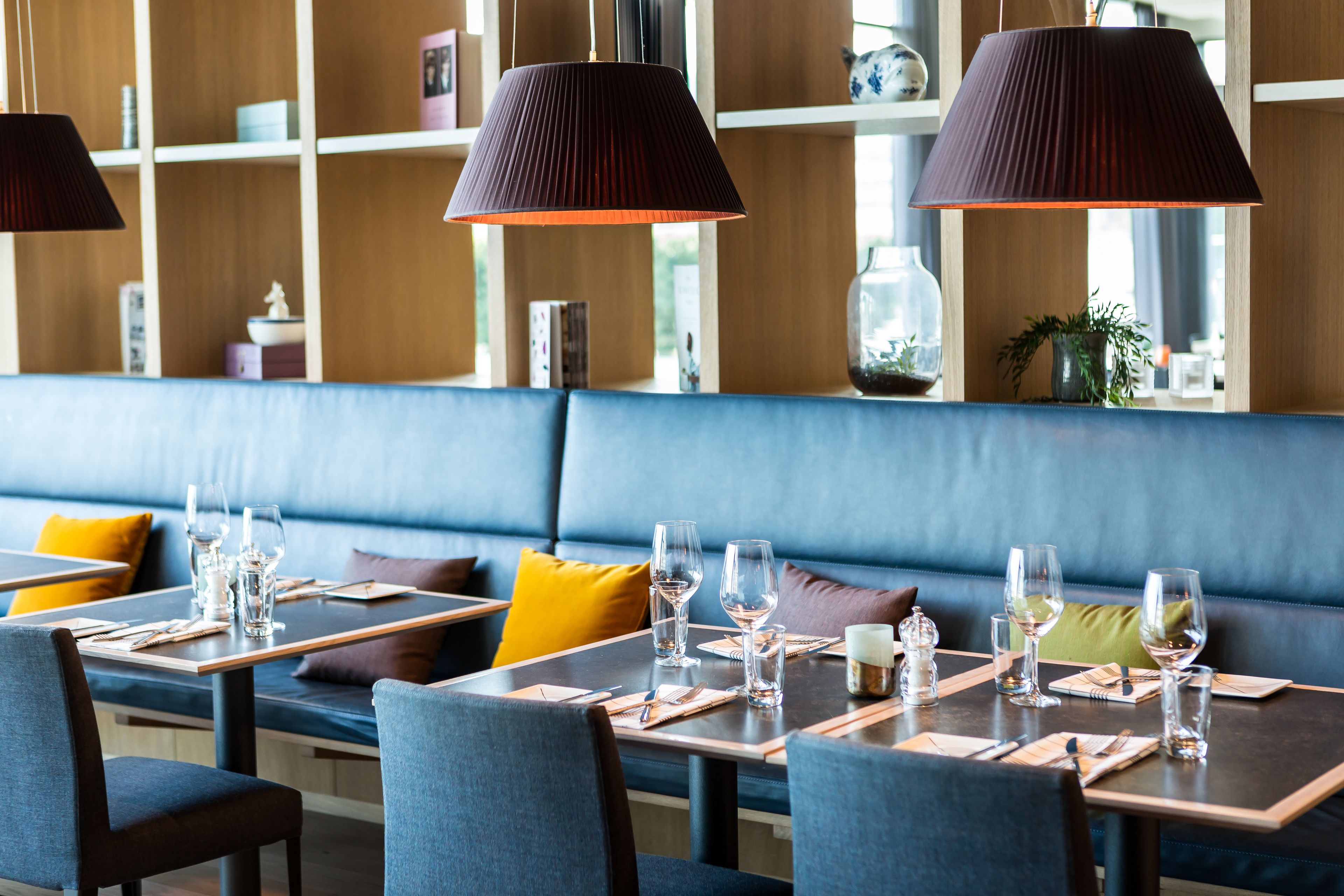 Een restaurant met houten planken, een blauwe met kussens bekleedde bank en tafels gedekt met wijnglazen en bestek.