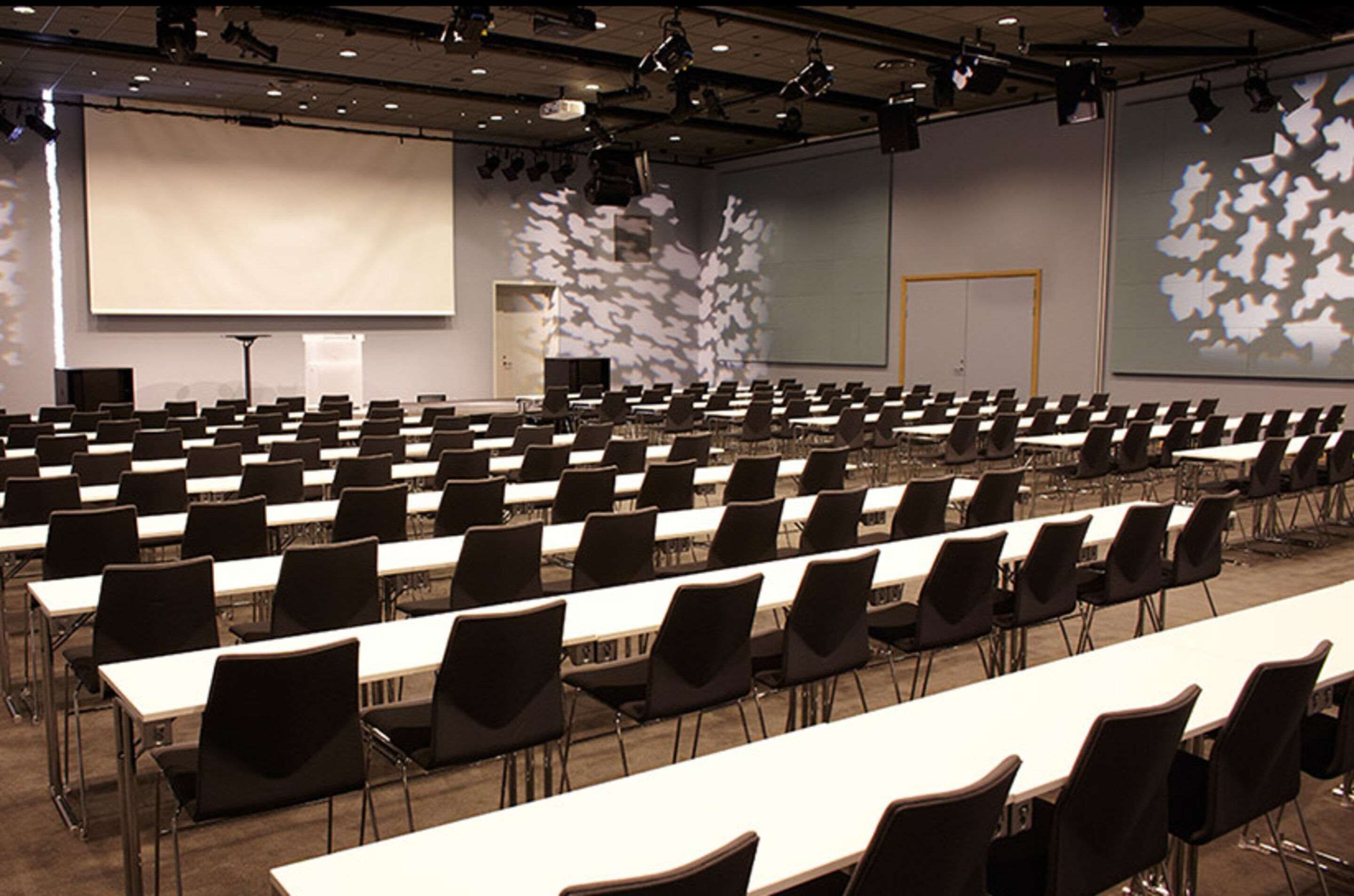Une salle de conférence avec des rangées de tables et de chaises, un grand écran et un écran de projection.