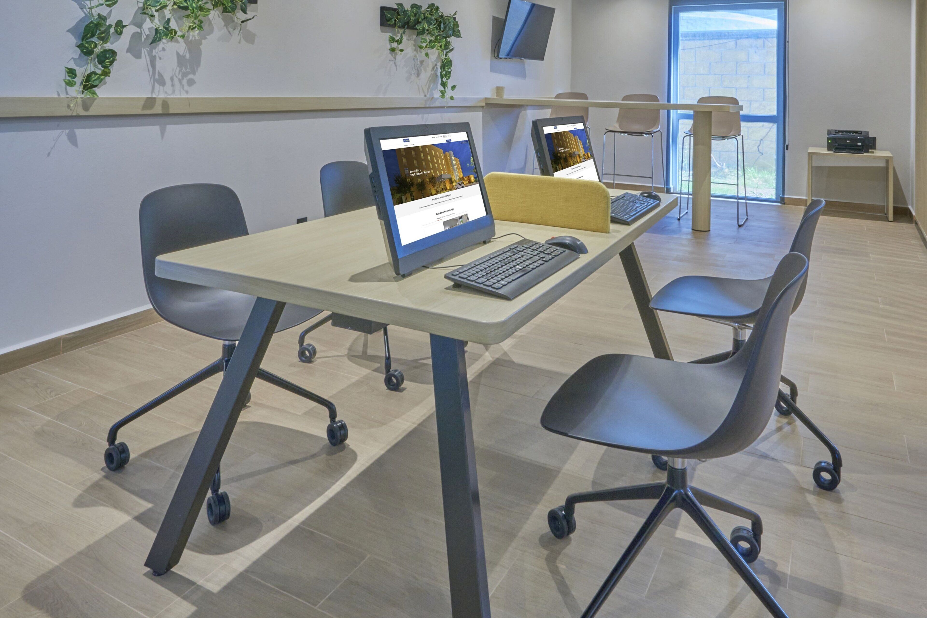 Een moderne kantoorruimte met twee bureaus, elk met een computermonitor, toetsenbord en muis. Er zijn vier stoelen, één bij elk bureau. De ruimte heeft licht houten vloeren, een groot raam en een aan de muur bevestigde televisie.