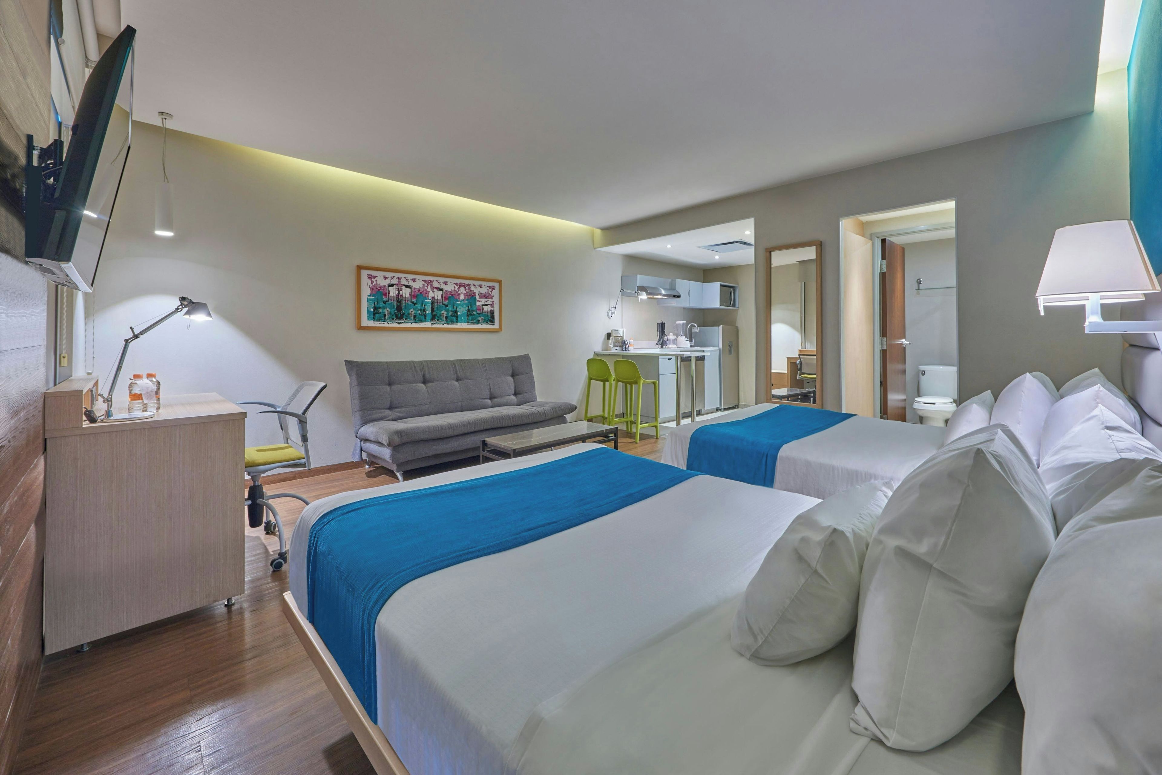 Habitación de hotel con dos camas, un sofá, un escritorio con lámpara, una kitchenette pequeña y un baño visible a través de una puerta abierta.