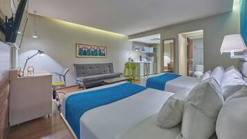 Habitación de hotel con dos camas, un sofá, un escritorio con lámpara, una kitchenette pequeña y un baño visible a través de una puerta abierta.