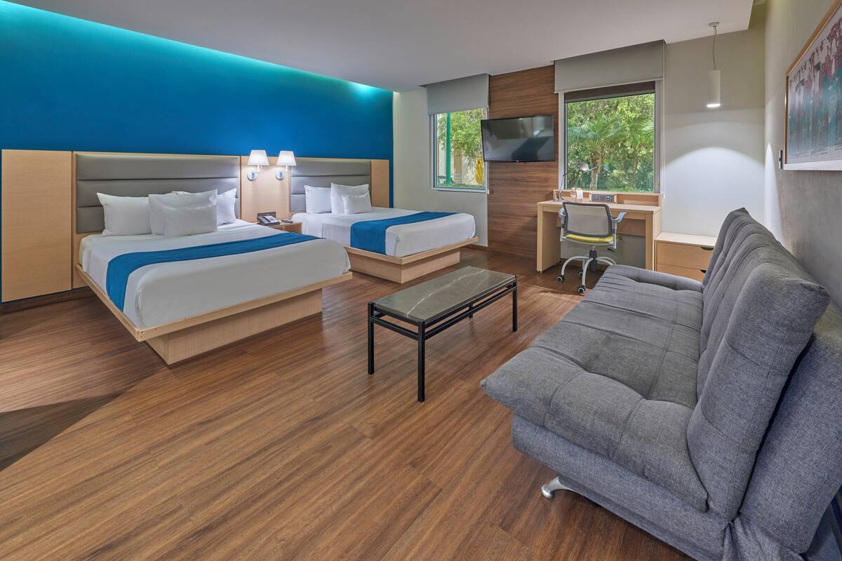 Habitación de hotel con dos camas, un escritorio, un sofá gris y un televisor.