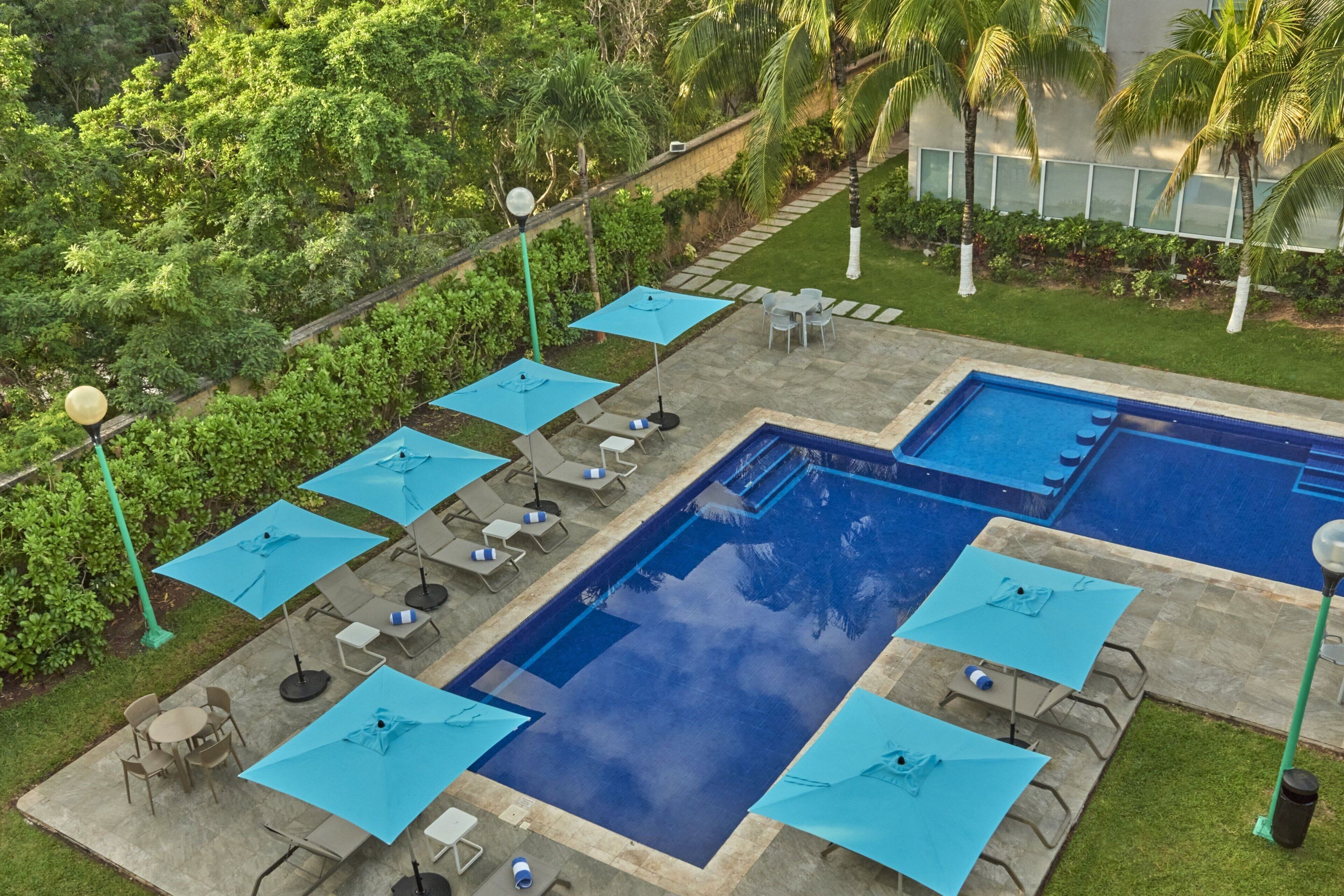 A swimming pool with blue umbrellas and lounge chairs, surrounded by greenery and a stone pathway.