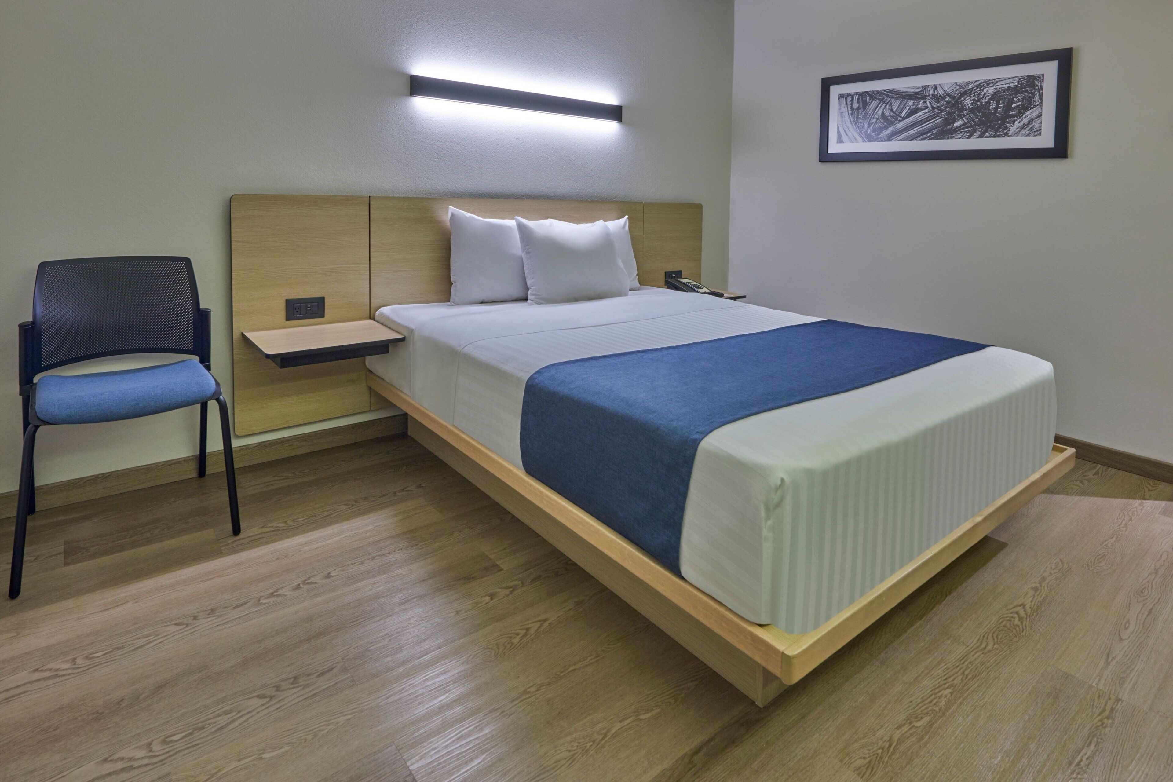 A hotel room with a bed, a chair, a bedside table, and a wall-mounted light fixture.