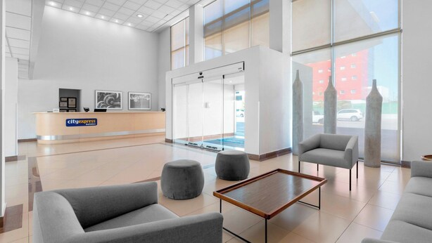 Un hall d’hôtel moderne avec un comptoir d’accueil, des sièges gris et de grandes fenêtres.
