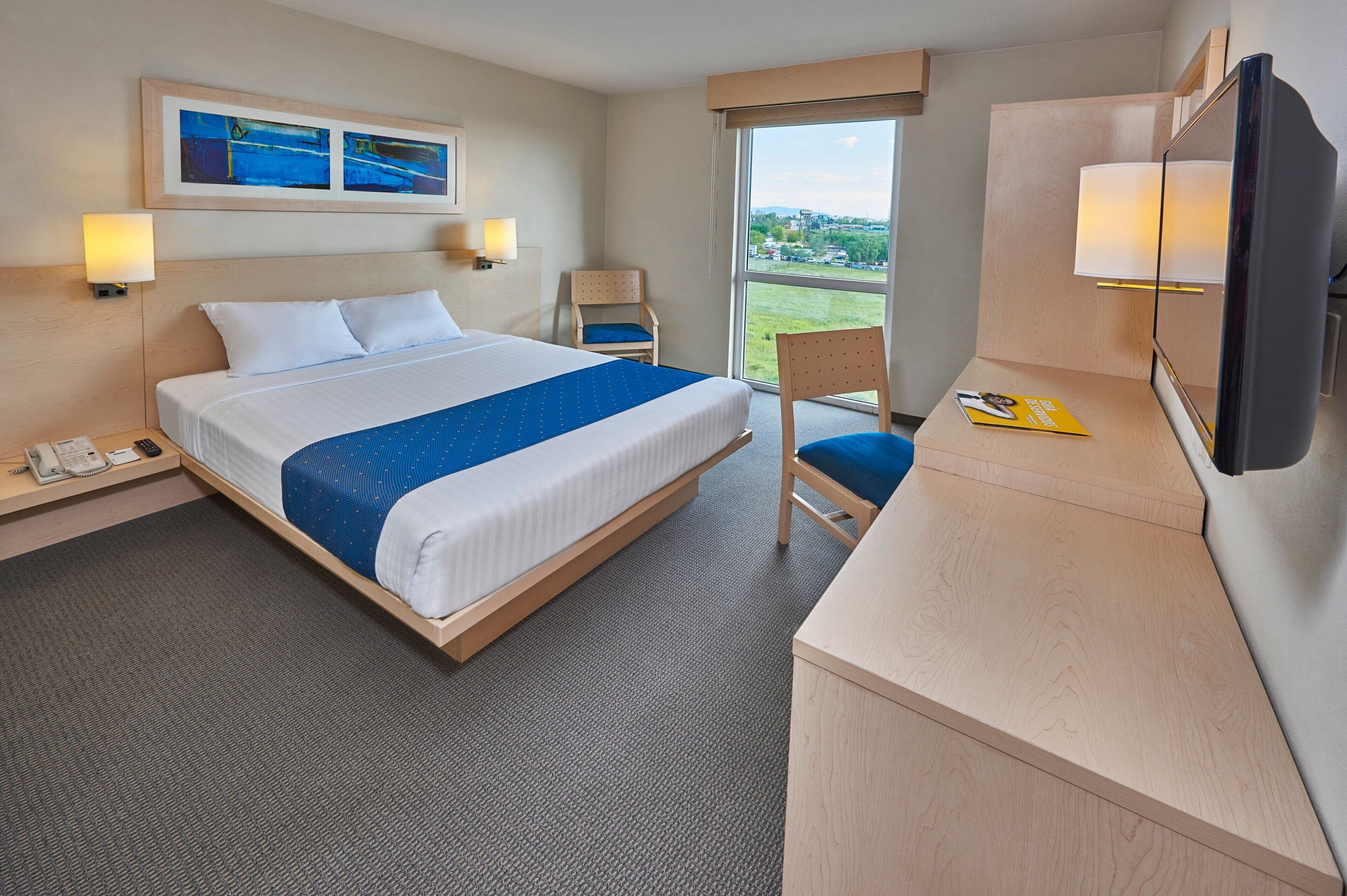 A hotel room with a bed, desk, chair, and large window offering a view.
