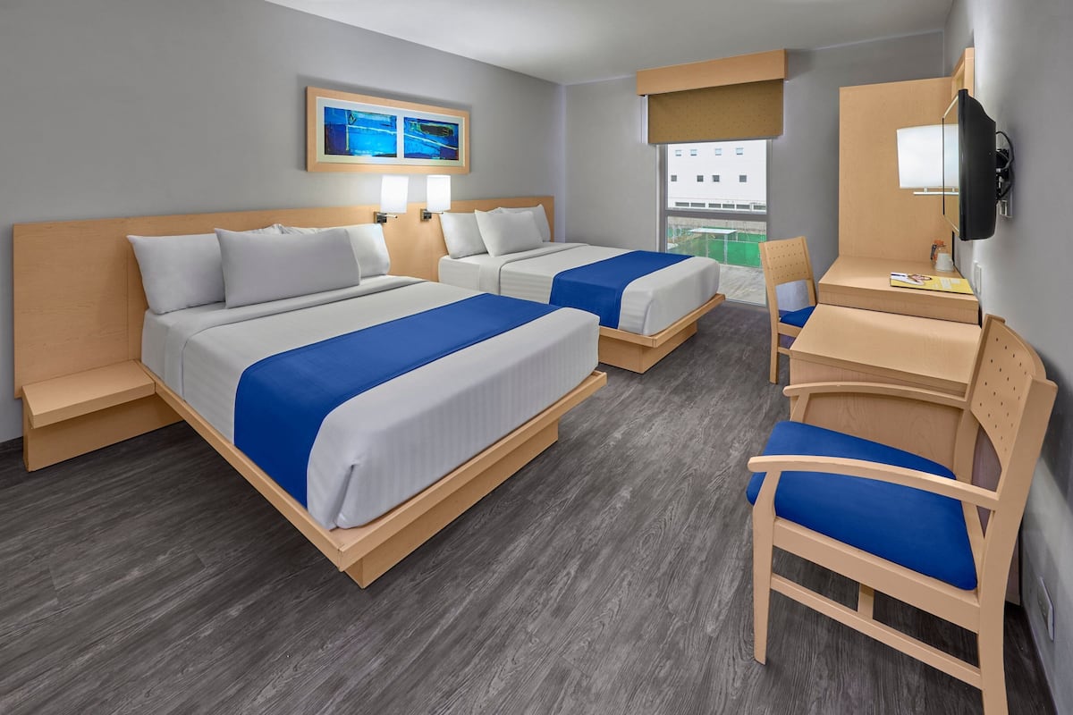 Habitación de hotel con dos camas, un escritorio, una silla, un televisor y una ventana con vista a edificios.