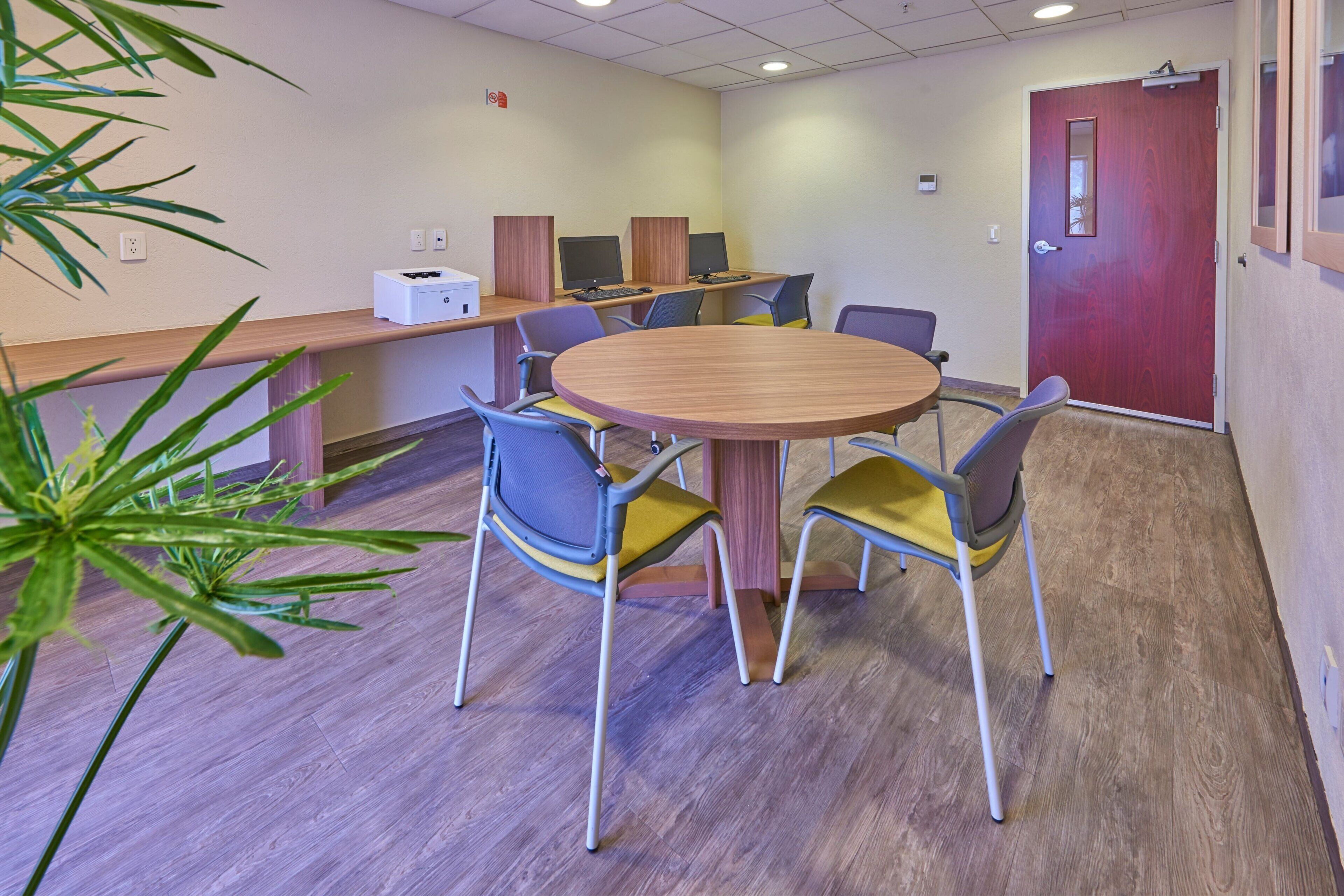 A conference room with a round table, four chairs, a computer station with two monitors, and a red door.