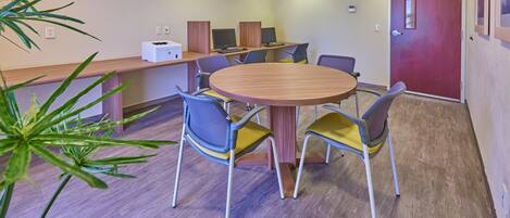 A conference room with a round table, four chairs, a computer station with two monitors, and a red door.