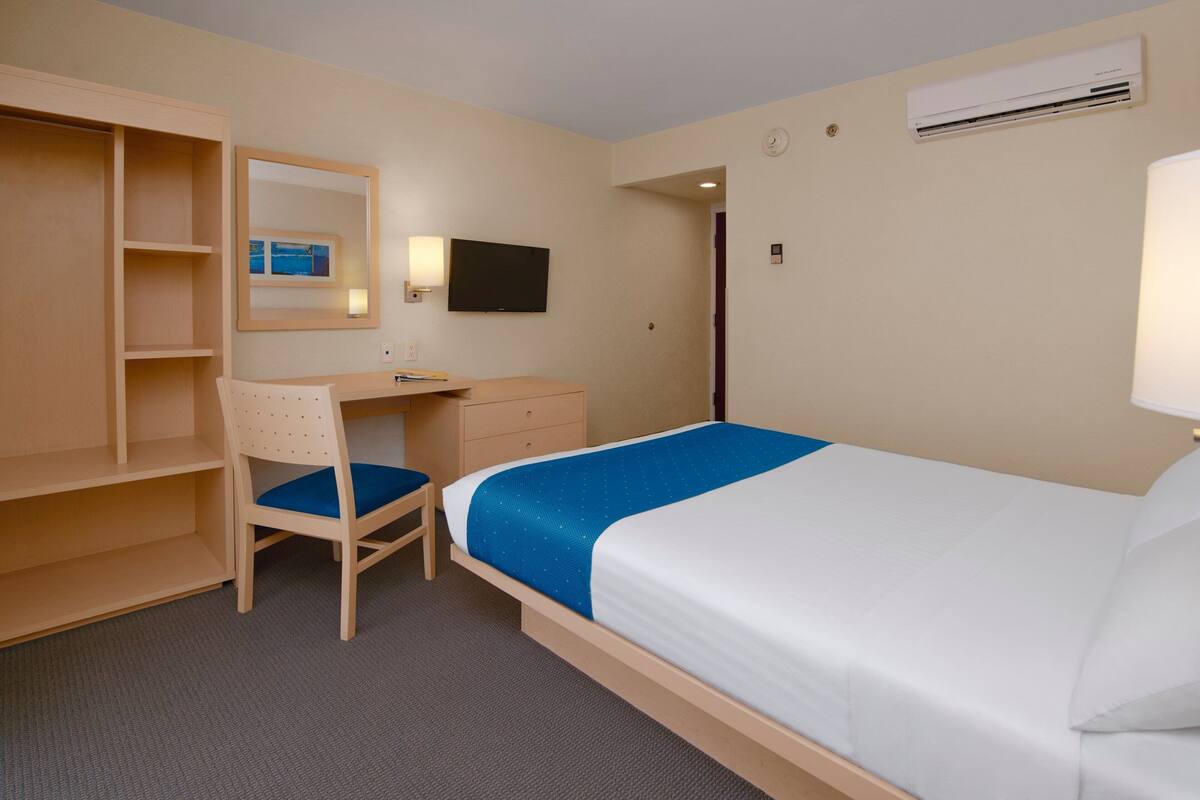 A hotel room with a bed, a desk, a chair, a wardrobe, a TV, and an air conditioning unit.