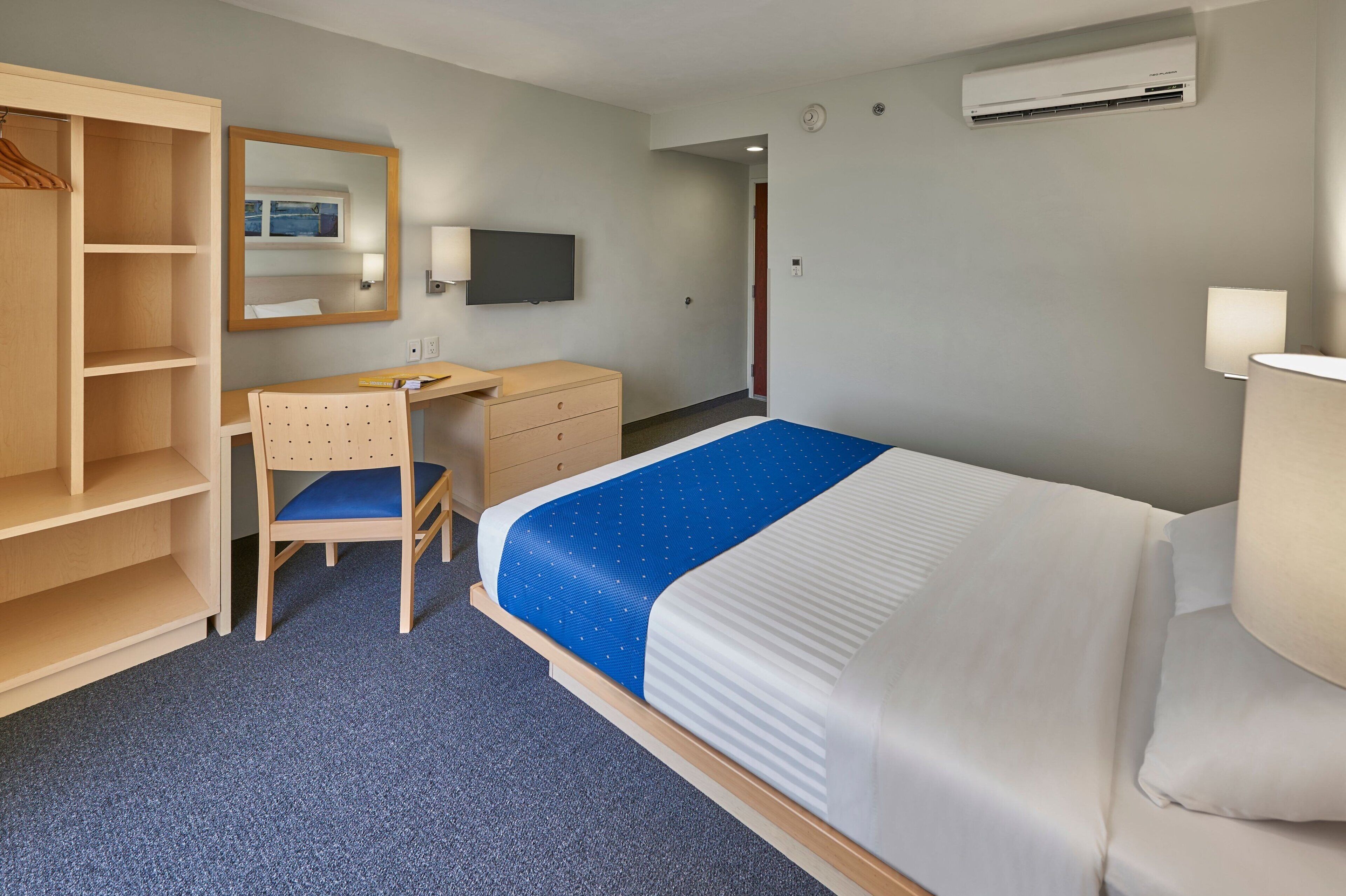 A hotel room with a bed, a desk, a chair, a wardrobe, a mirror, a television, and an air conditioning unit.