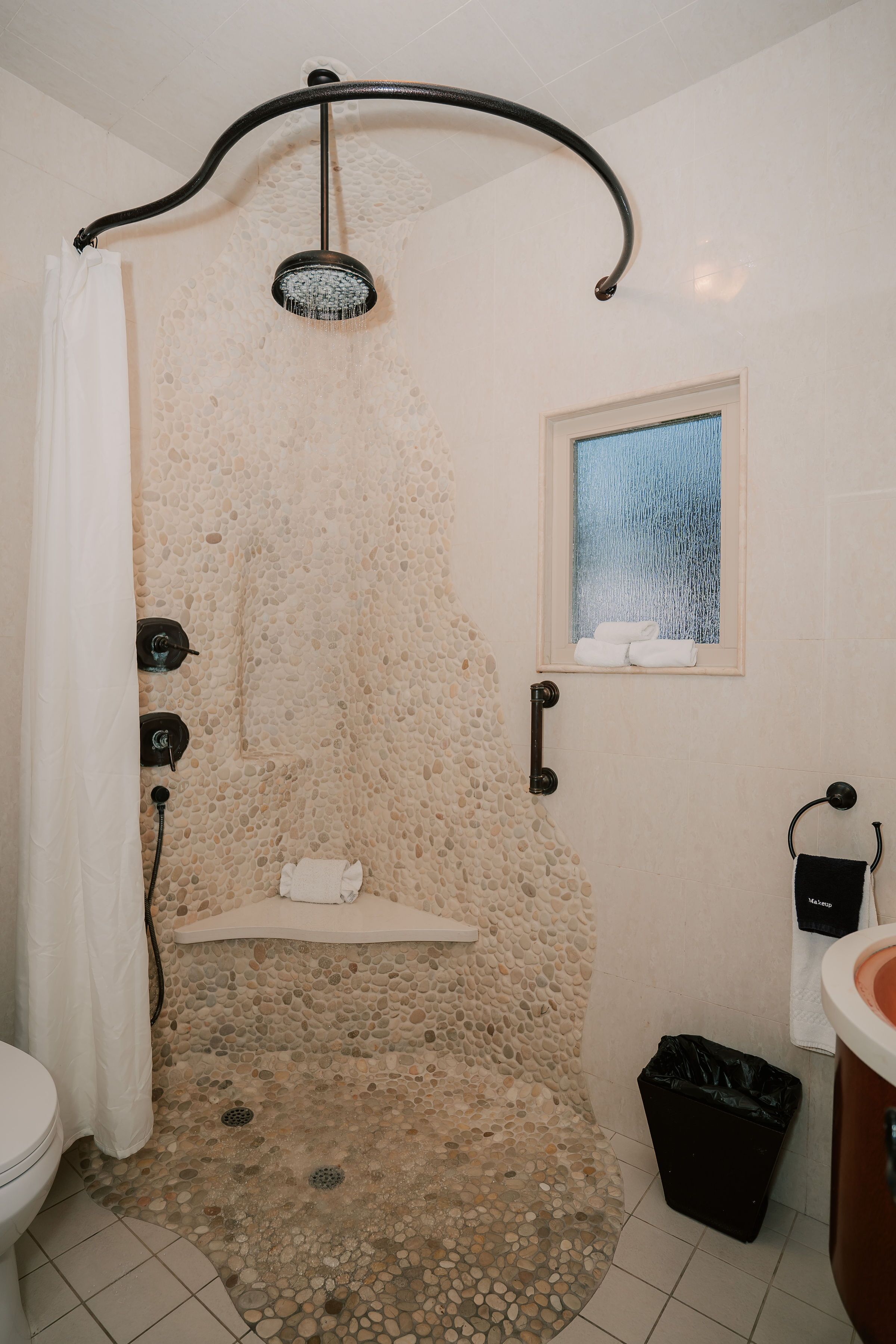 A stone-walled shower with a rain showerhead, a small window with a towel hanging, and a black trash bin.