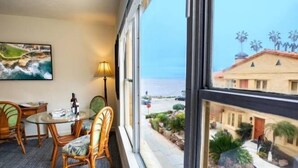 A room with a dining table and chairs, a painting, a lamp, and a view of the ocean and a building with palm trees. - Pantai Inn (La Jolla)