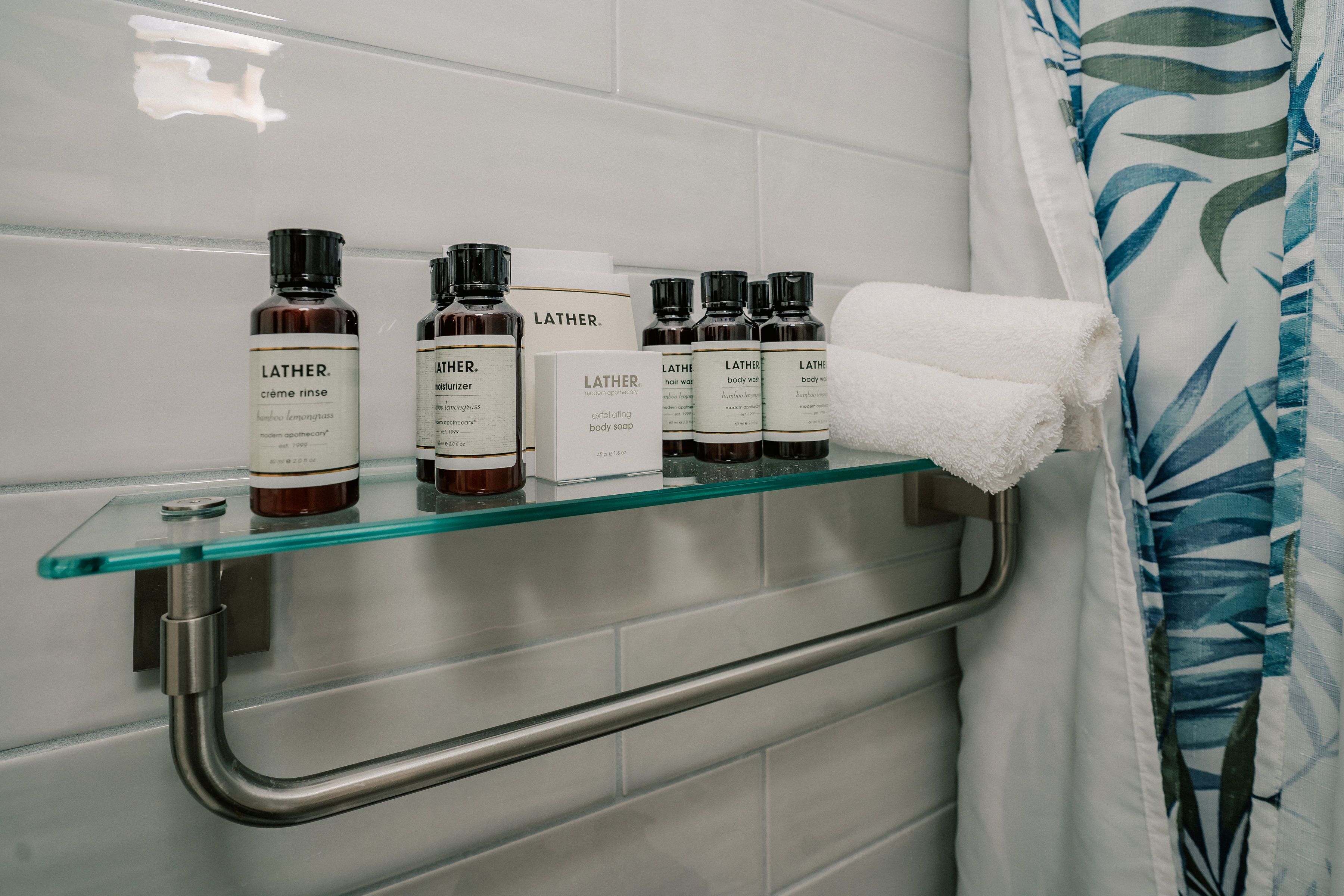 Une étagère dans la salle de bain avec des flacons de gel douche et d’après-shampoing de la marque Lather, une serviette blanche et un receveur de douche en verre.
