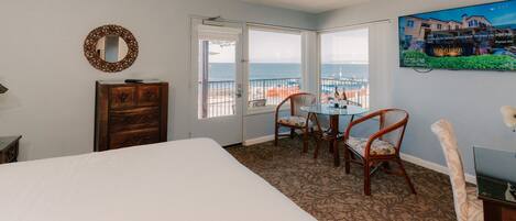 A hotel room with a bed, a dresser, a desk with chairs, a large window with a view of the sea, and a painting on the wall.