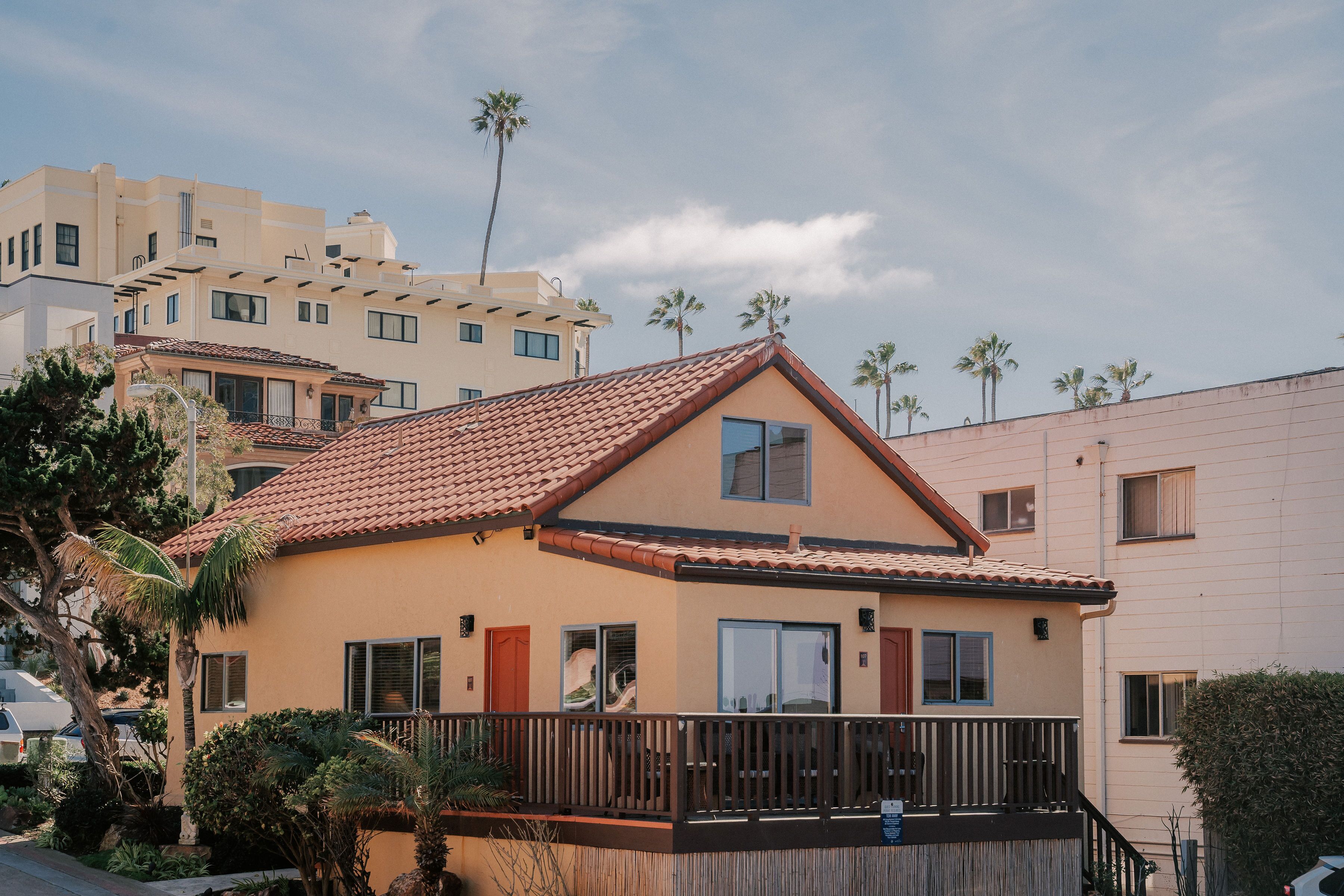 A two-story house with a red-tiled roof, a balcony with a wooden railing, and a small garden area with palm trees.