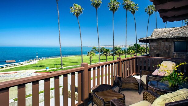 A balcony with wicker furniture, a stone building, and a view of the ocean and golf course. - Pantai Inn (La Jolla)