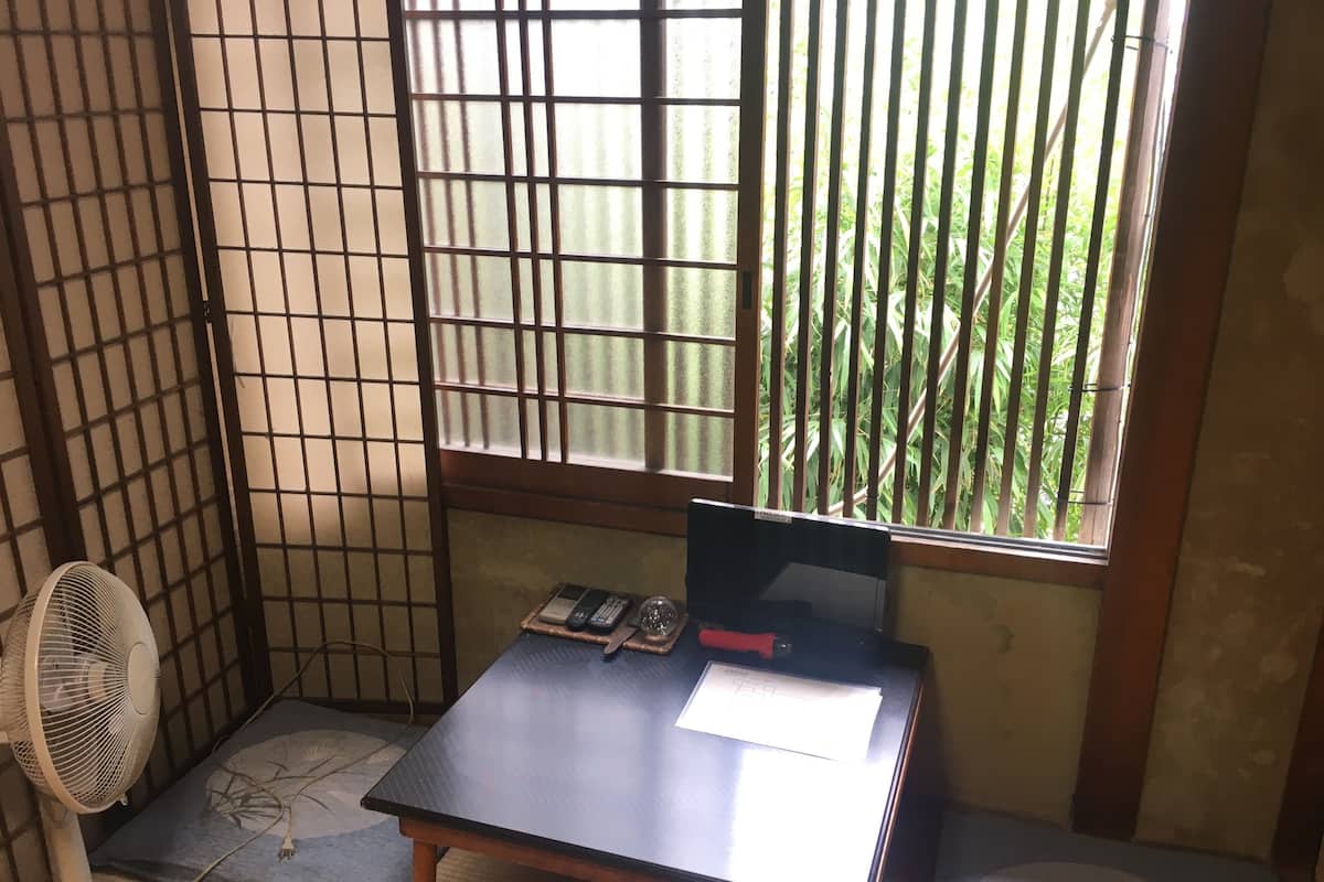 Una habitación con suelo de tatami, un escritorio con computadora, un ventilador y una ventana con bambú afuera.