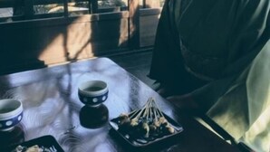 Une personne vêtue d’un costume traditionnel est assise à une table devant un plateau de nourriture et deux tasses.