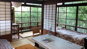 Une pièce de style japonais traditionnel, avec un sol recouvert de tatamis, une petite table recouverte d'une nappe en dentelle, un lit agrémenté d'une couvre-lit à motifs floraux et de grandes fenêtres offrant une vue sur la verdure.