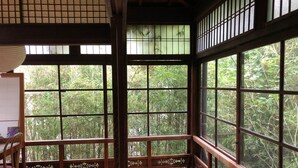 Une pièce de style japonais traditionnel, avec un sol recouvert de tatamis, une basse table en bois et un banc. De grandes fenêtres à cadre en bois offrent une vue sur une végétation luxuriante.