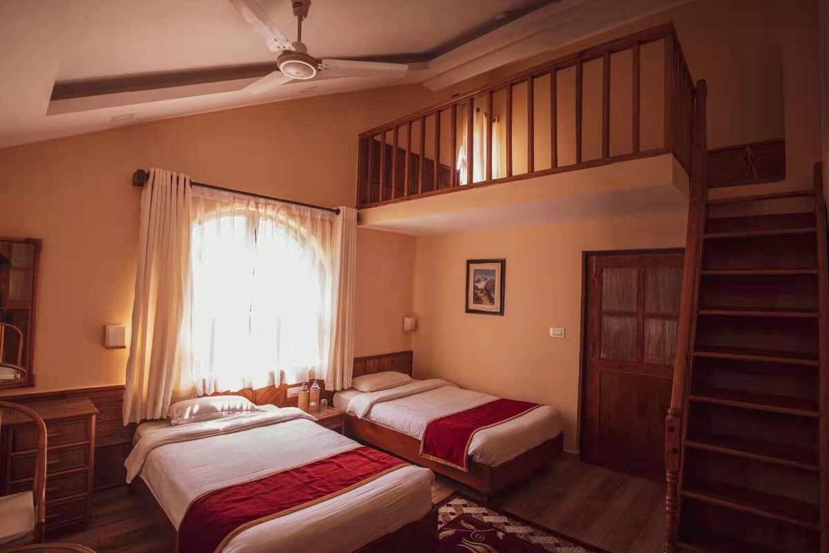 Una habitación con dos camas, una escalera de madera, un ventilador de techo y una ventana con cortinas.